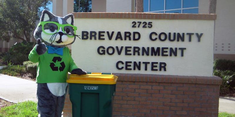 RC the Recycling Cat standing in front of the Brevard County Government Center building with a recycling trash bin. 